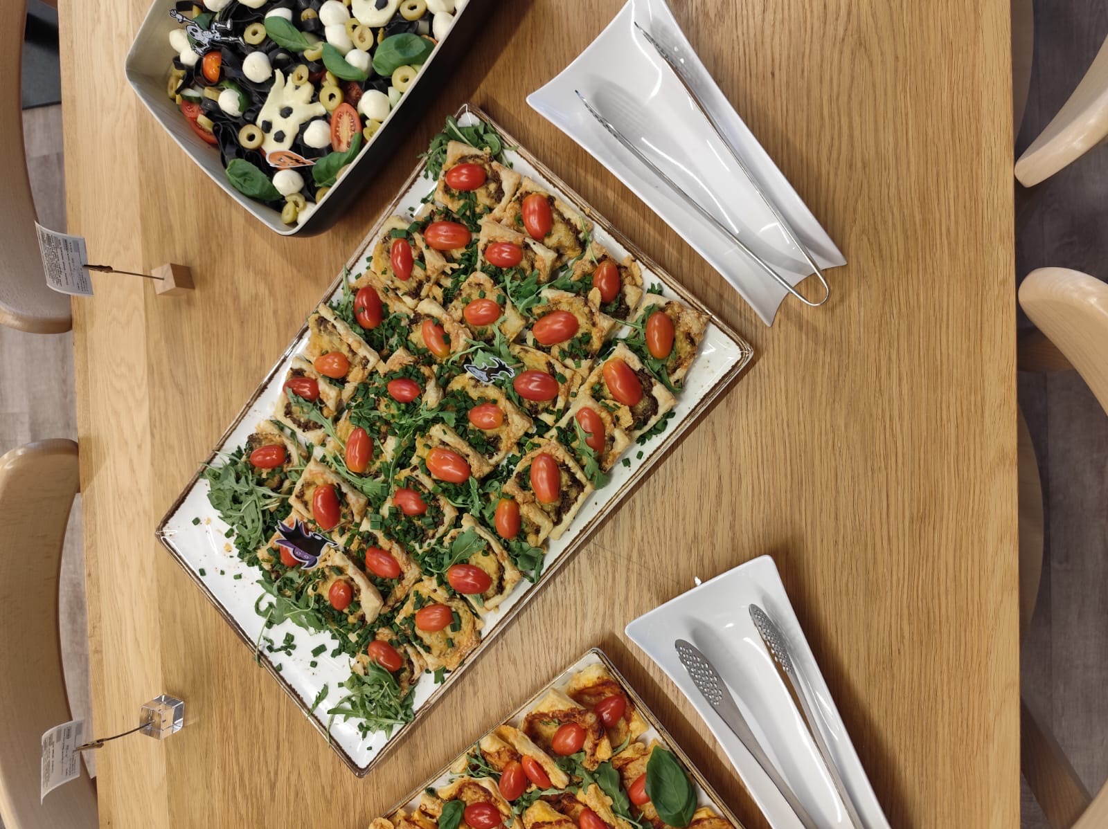 Widok z góry na stół z cateringiem: kwadratowe tartaletki z pomidorkami cherry i zieleniną, sałatka z czarnym makaronem i mozzarella, szczypce do nakładania.