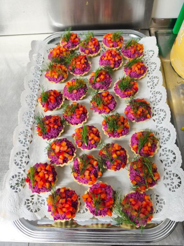 Tarty z buraczkową pastą i czerwoną papryką, udekorowane koperkiem, ułożone na srebrnej tacy z serwetką. Widoczne metalowe elementy wyposażenia kuchni w tle.