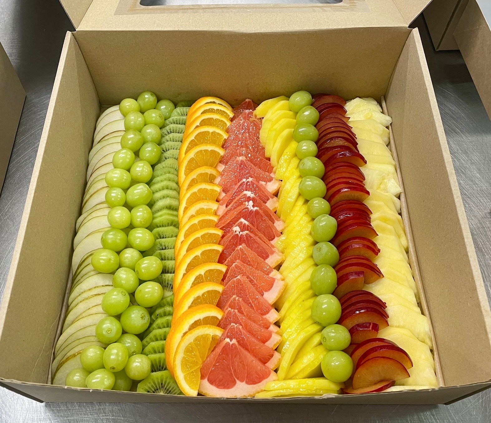 Elegancka kartonowa taca z owocami: jabłka, winogrona, kiwi, pomarańcze, grejpfruty, mango, śliwki i ananas, ułożone w rzędach, idealne na catering.