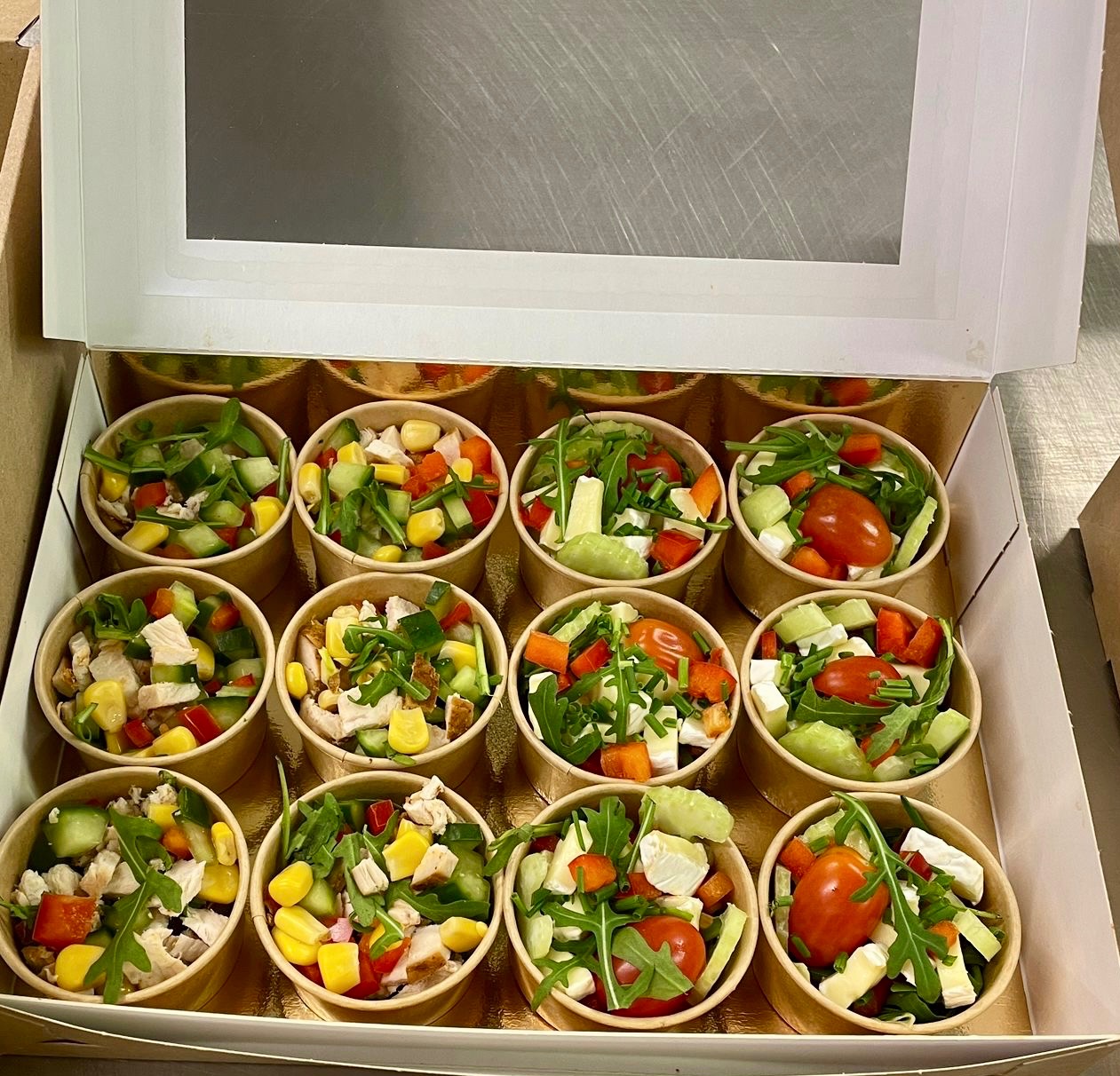 Zestaw 12 porcji sałatek w papierowych kubeczkach, ułożonych w pudełku z okienkiem. Różne kompozycje: kurczak, kukurydza, pomidor, ogórek, rukola, ser.