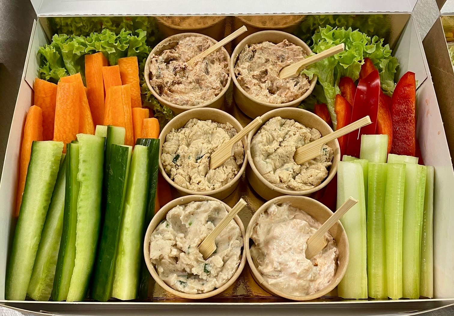 Elegancki box cateringowy z warzywami (marchew, seler naciowy, ogórek, papryka) i dipami w papierowych miseczkach z drewnianymi łyżeczkami, idealny na przyjęcie.