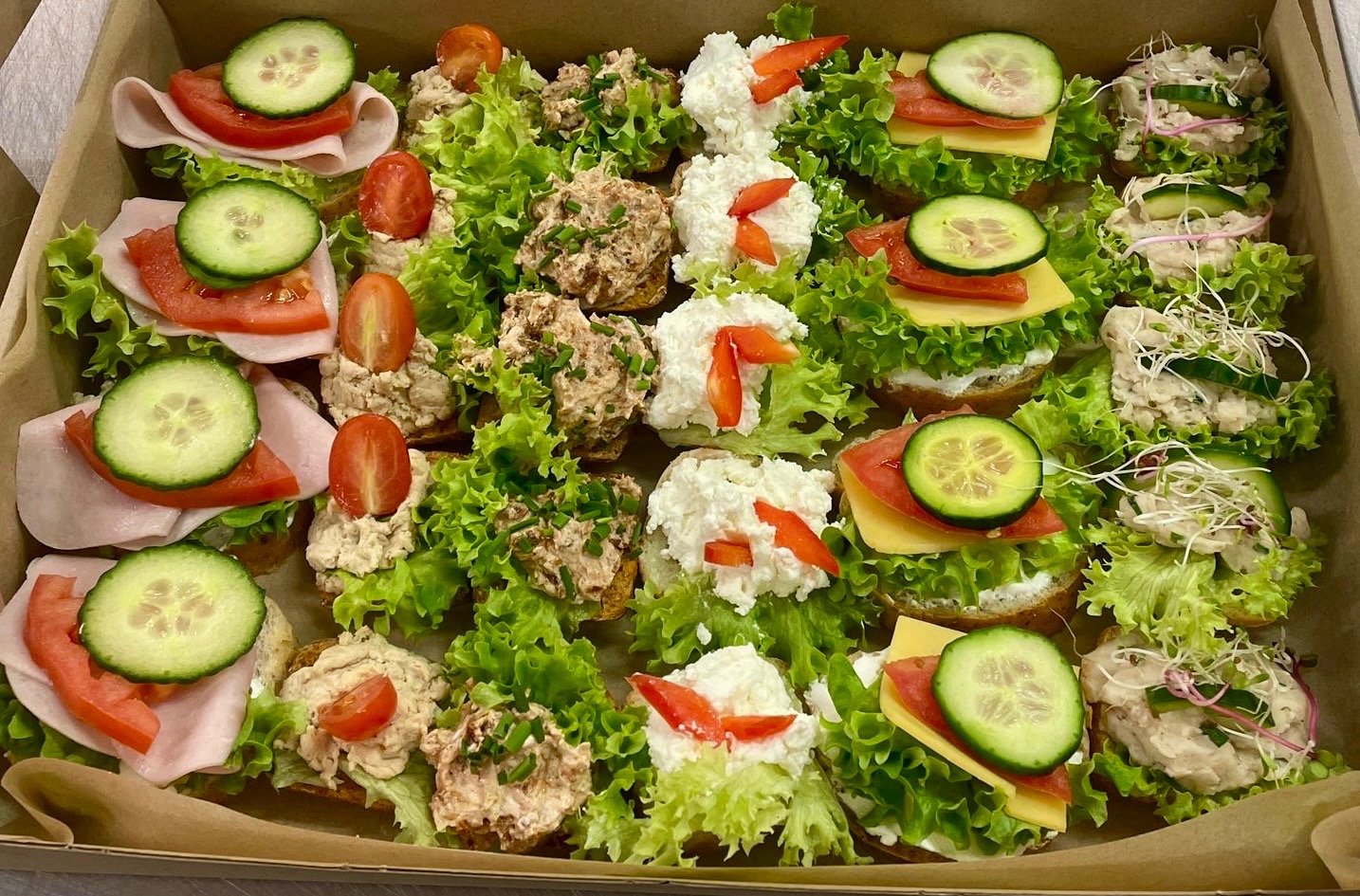 Różnorodne kanapki na pieczywie, z sałatą, pomidorem, ogórkiem, szynką, serem i pastami, ułożone w kartonowym pudełku. Catering na imprezę.