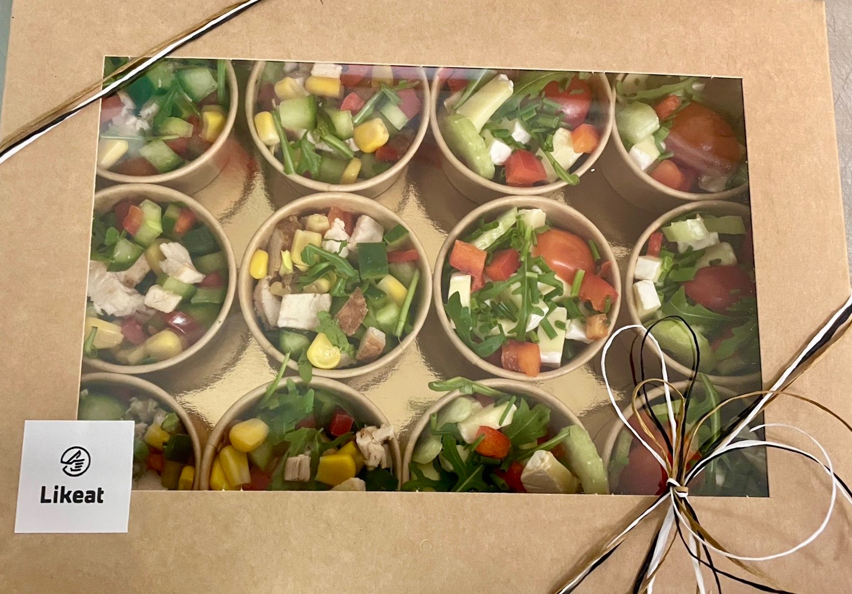 Zestaw 12 porcji sałatek w papierowych kubeczkach, zapakowanych w kartonowe pudełko z okienkiem, przewiązane sznurkiem. Różne kompozycje warzyw i dodatków.