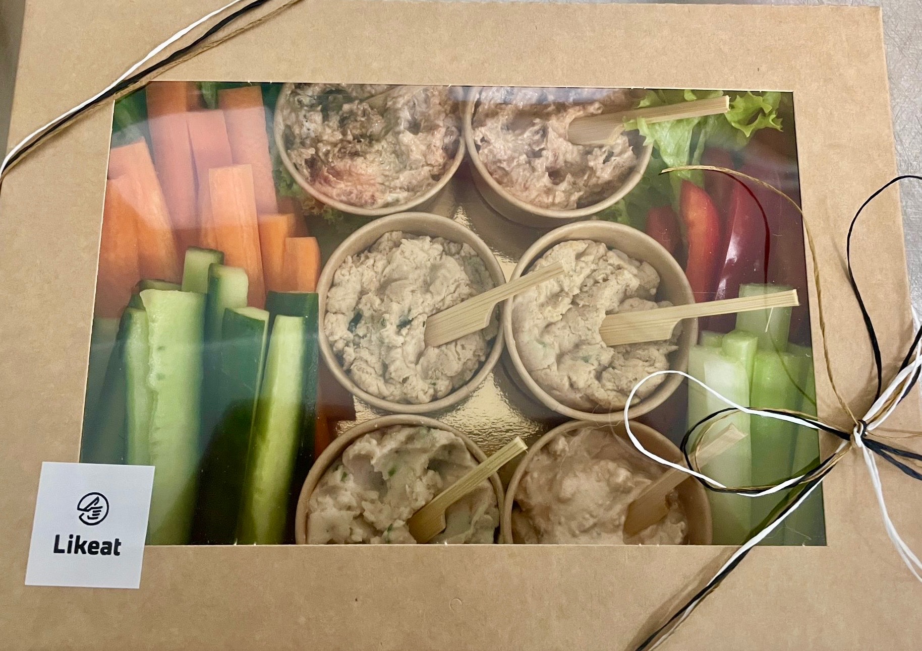 Elegancki box cateringowy z warzywami (marchew, ogórek, papryka) i dipami w papierowych miseczkach, ozdobiony sznurkiem. Widoczne logo Likeat.