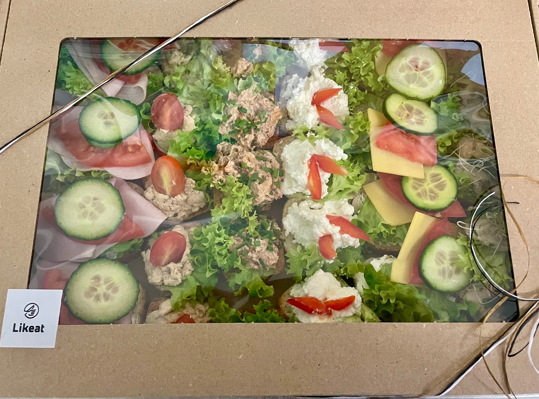Zestaw kanapek w pudełku cateringowym z logiem Likeat. Kanapki z różnymi dodatkami: ogórek, pomidor, ser, szynka, sałata, pasta.