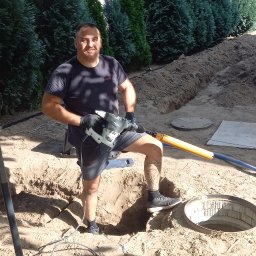 Arkoinstal - Uśmiechnięty instalator z młotem udarowym stoi w wykopie obok studzienki kanalizacyjnej na tle zielonych drzew. Widoczny kabel i szpadel.