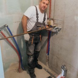 Arkoinstal - Hydraulik w roboczym kombinezonie, z poziomicą, montuje instalację wodną w surowym wnętrzu. Widoczne narzędzia i rury.