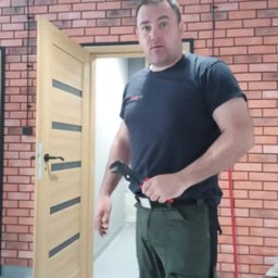 Arkoinstal - Hydraulik z kluczem nastawnym na tle ceglanej ściany i otwartych drzwi. Widoczne przewody elektryczne. Ujęcie z bliska, na pierwszym planie narzędzie.