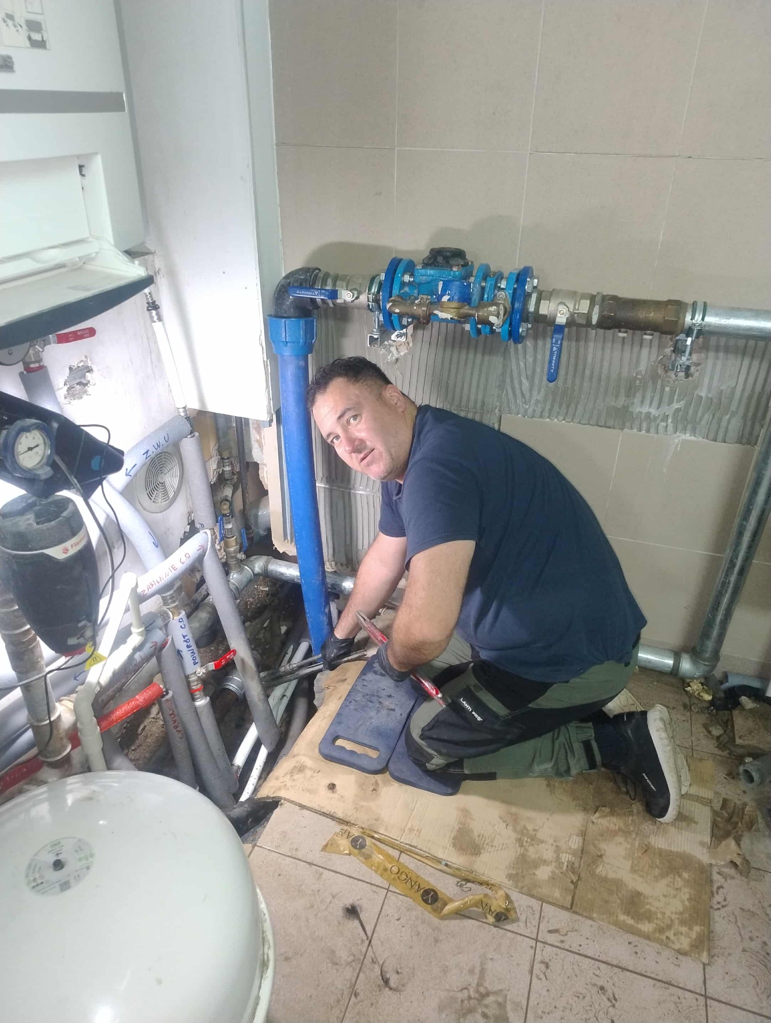 Hydraulik w trakcie pracy nad instalacją wodną w pomieszczeniu z beżowymi płytkami. Widoczne rury, zawory i narzędzia. Fachowiec klęczy na podłodze, skupiony na zadaniu.