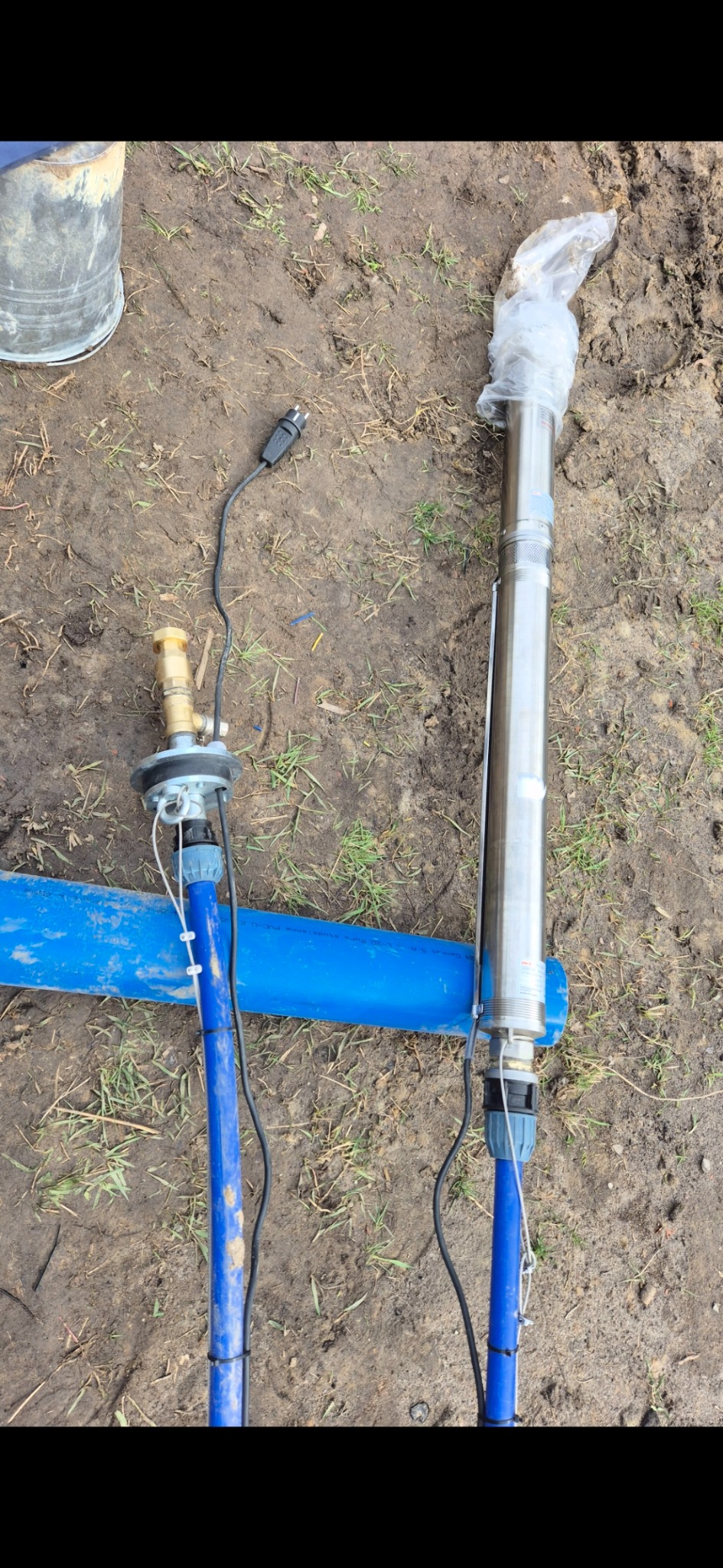 Dwie pompy głębinowe: jedna w folii, druga z osprzętem, leżą na niebieskiej rurze na ziemi. Widoczny kabel zasilający i wiadro w tle.