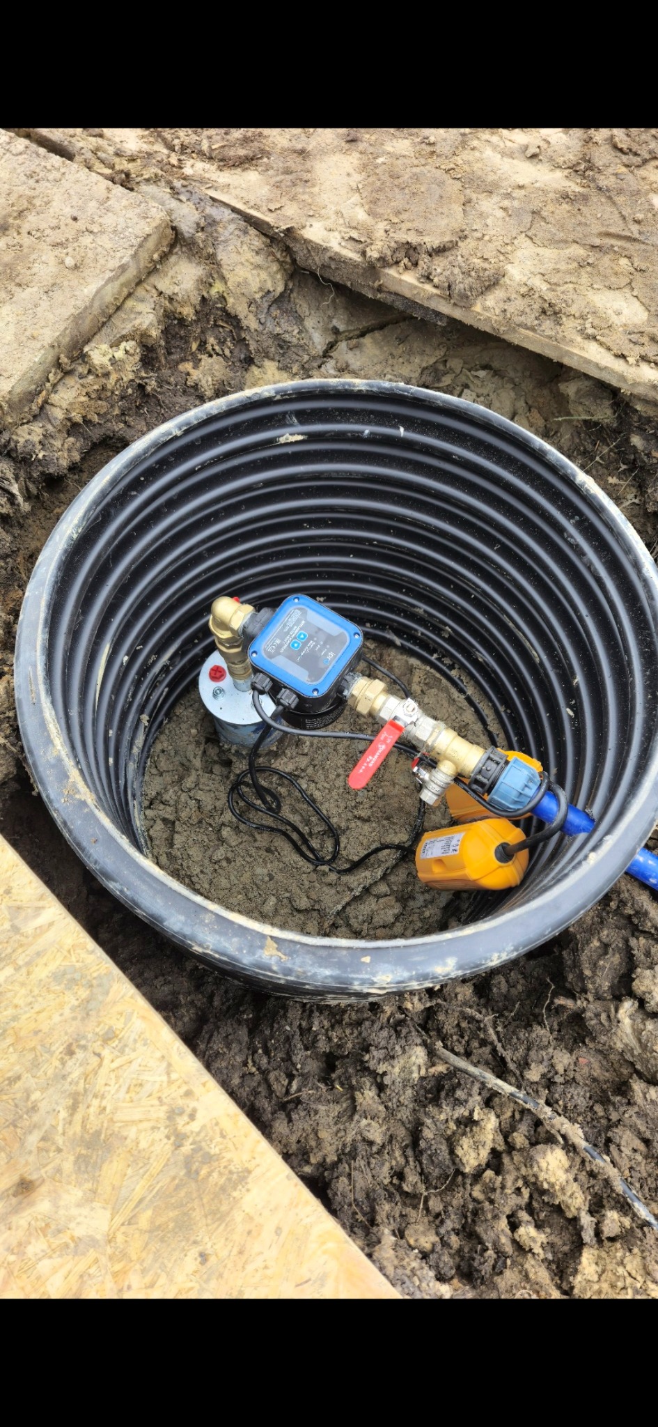 Studnia głębinowa z zamontowaną pompą i osprzętem w czarnej rurze karbowanej, widok z góry na ziemiste tło.