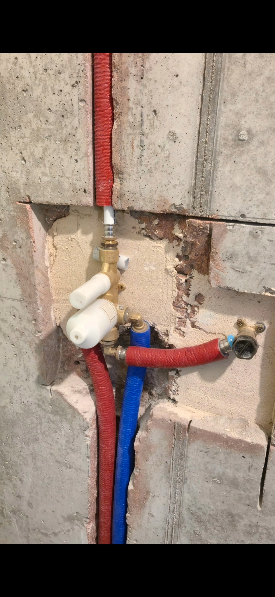 Fragment ściany z widoczną instalacją wodną: rury w izolacji (czerwona), rura niebieska, zawór z białymi pokrętłami. Widoczne ślady po kuciu w betonie.