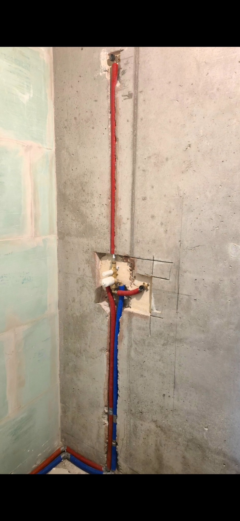 Instalacja wodna w łazience w trakcie montażu: czerwone i niebieskie rury prowadzone wzdłuż surowej, betonowej ściany, z widocznymi przyłączami armatury.