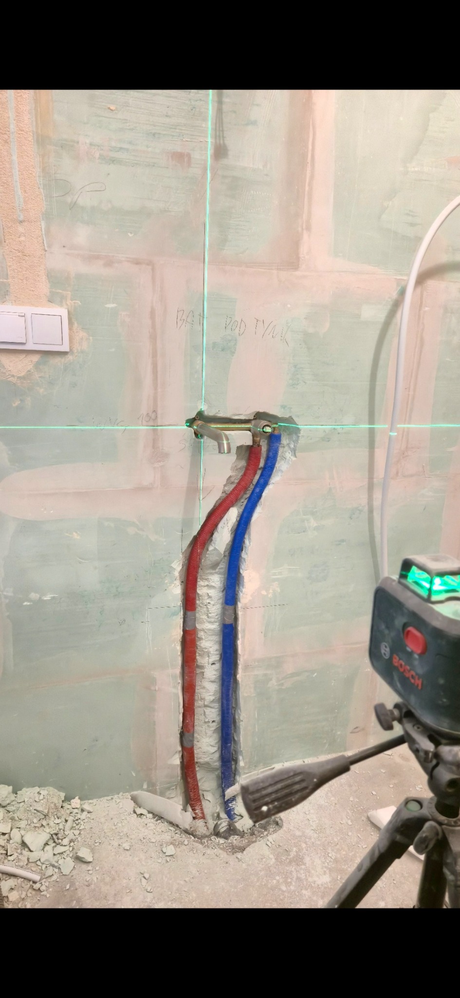 Montaż rur wodnych (czerwona i niebieska) w ścianie, z użyciem poziomicy laserowej. Widoczne ślady po kuciu w ścianie i gruz na podłodze.