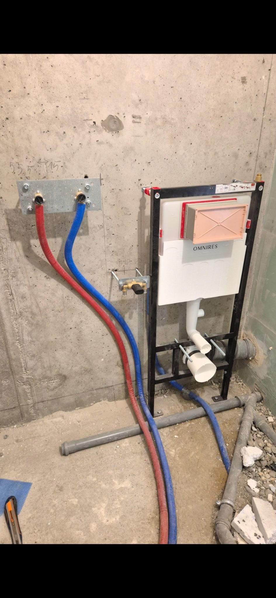 Stelaż podtynkowy WC marki OmniRes z podłączonymi rurami doprowadzającymi wodę (czerwona i niebieska) na tle surowej ściany betonowej w trakcie montażu.