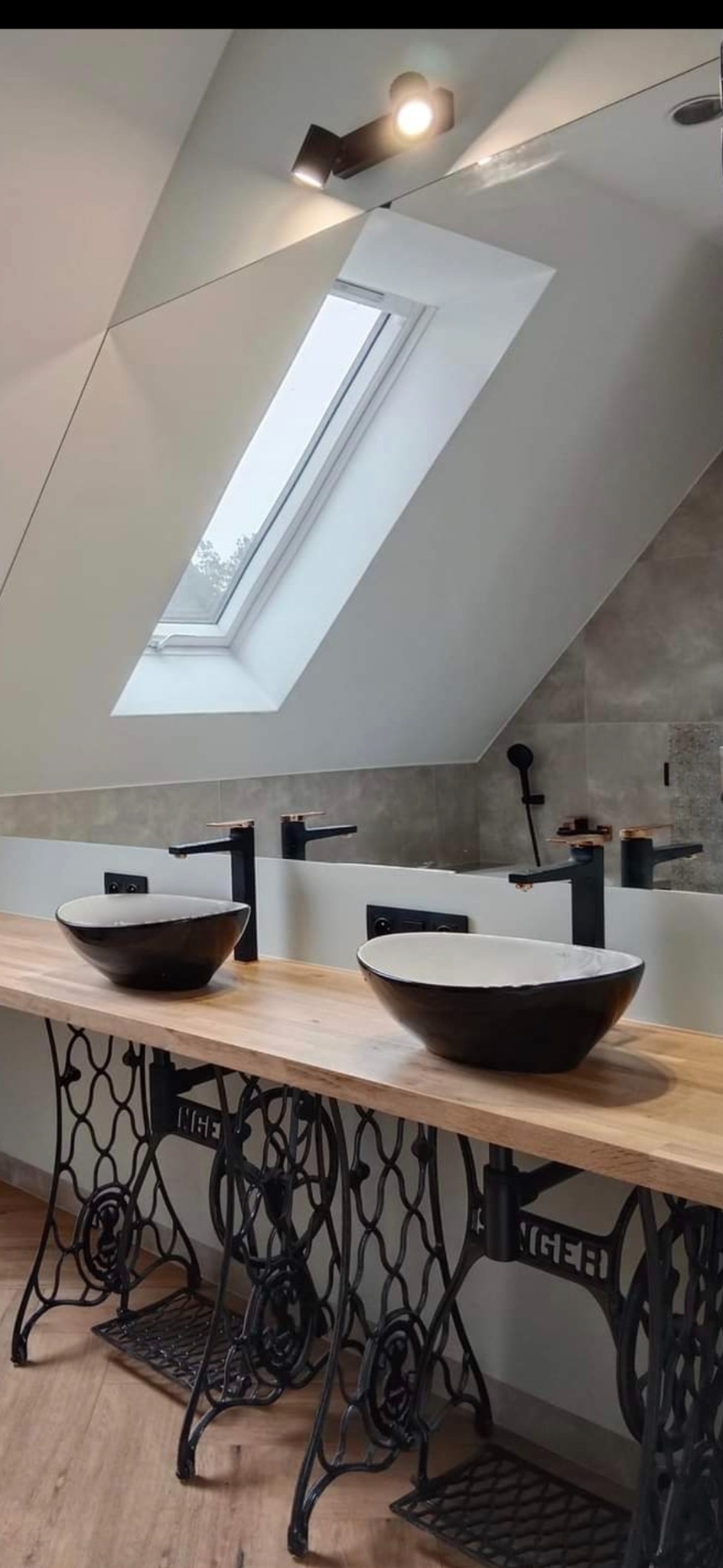 Nowoczesna łazienka na poddaszu z umywalkami w kształcie mis, czarną armaturą i blatem na podstawie maszyny do szycia Singer. Okno dachowe doświetla przestrzeń.