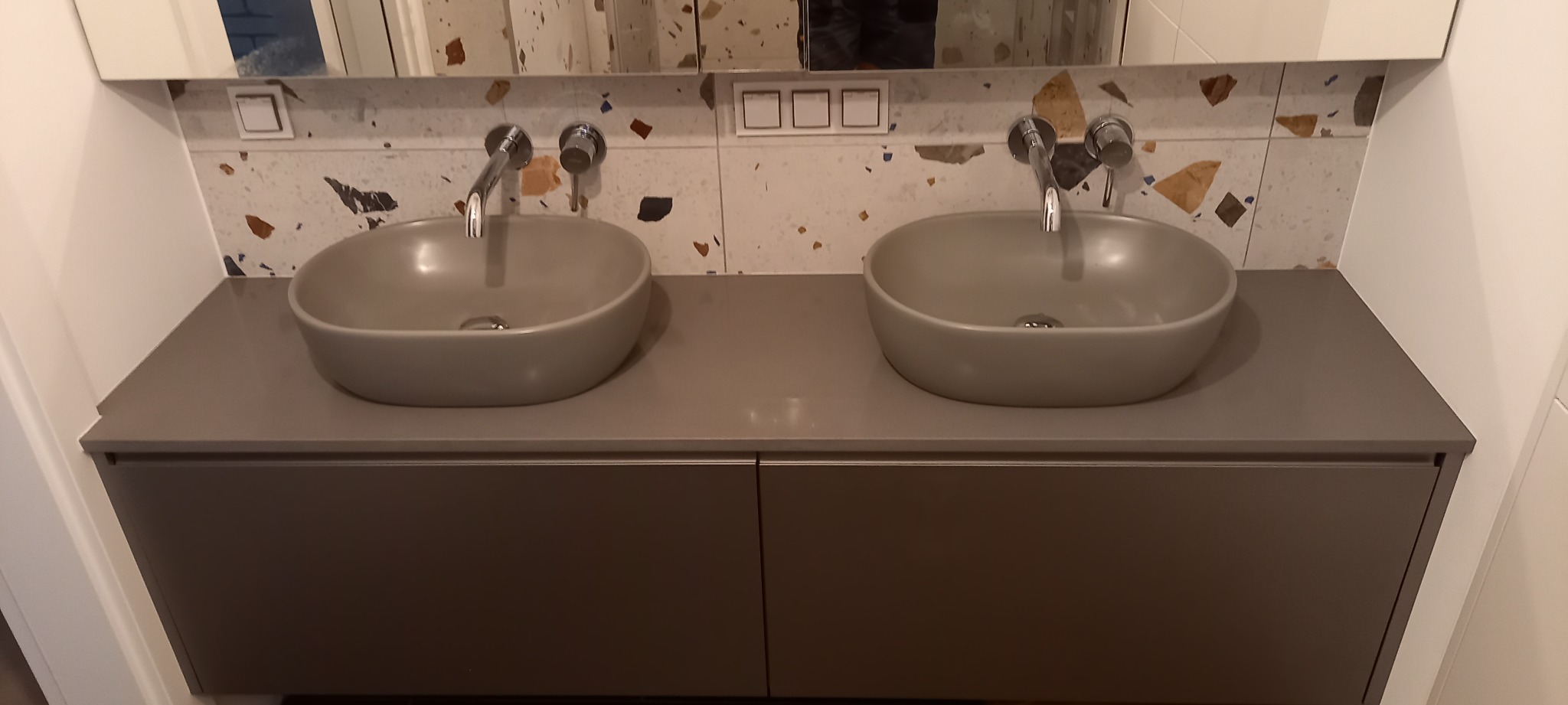 Dwie szare, owalne umywalki nablatowe z chromowanymi bateriami na szarym blacie z szafką w łazience z mozaiką. Nowoczesny design.