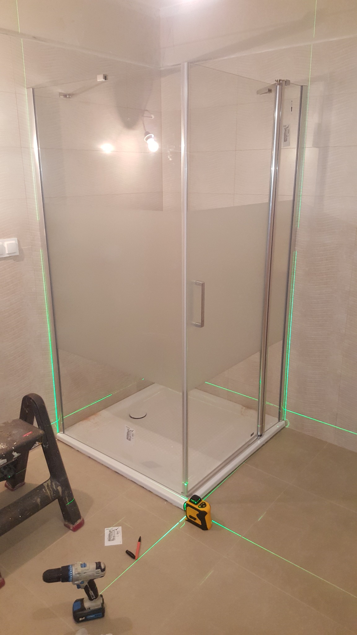 Montaż kabiny prysznicowej z użyciem poziomicy laserowej. Widoczna wiertarka, drabina i instrukcja. Ukończona instalacja w łazience z beżowymi płytkami.
