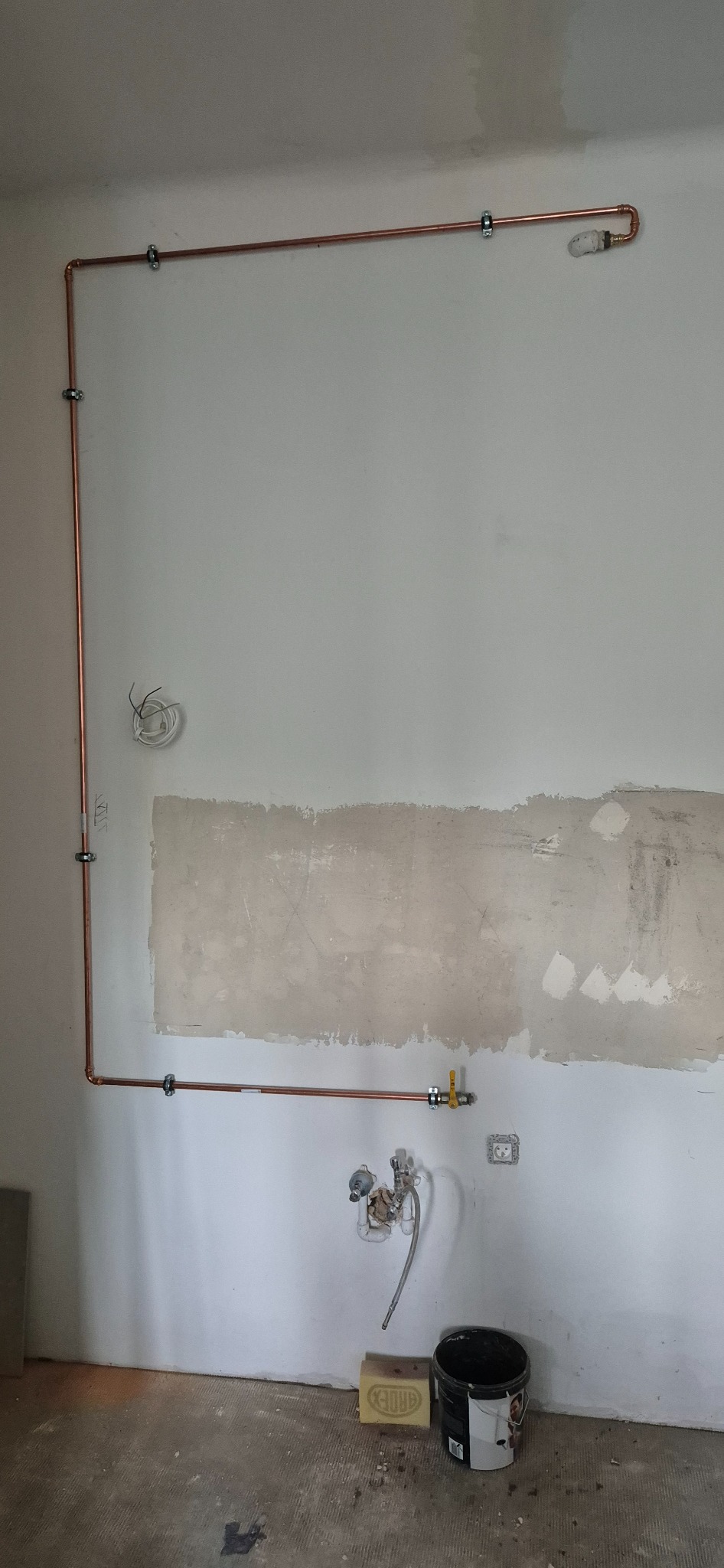 Miedziana instalacja gazowa na ścianie w trakcie remontu, widoczne rury, zawór i uchwyty mocujące. Ściana częściowo przygotowana do malowania.