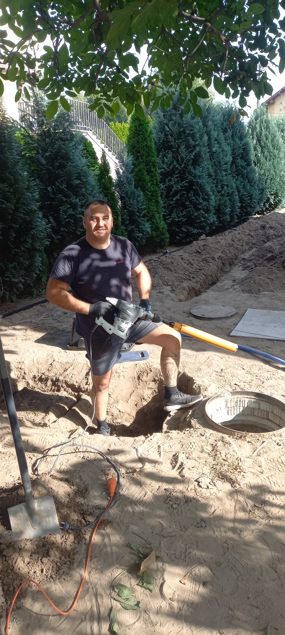 Uśmiechnięty instalator z młotem udarowym stoi w wykopie obok studzienki kanalizacyjnej na tle zielonych drzew. Widoczny kabel i szpadel.