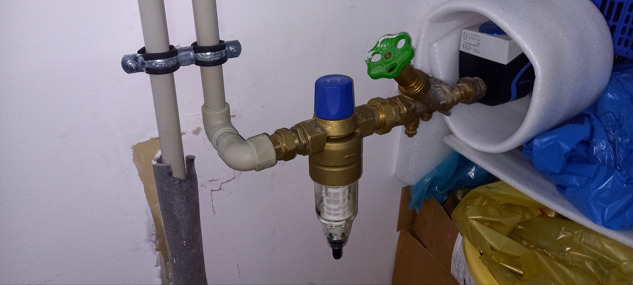 Zamontowany filtr wody z zaworem i rurami przy ścianie. Widoczne elementy instalacji hydraulicznej w pomieszczeniu.