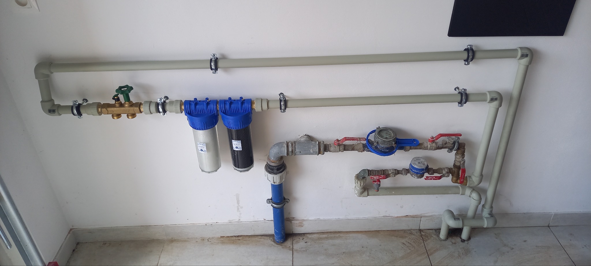 Kompleksowa instalacja wodna z filtrami i licznikami, rury prowadzone wzdłuż ściany, widoczne zawory i złączki. Szara kolorystyka rur kontrastuje z niebieskimi filtrami.
