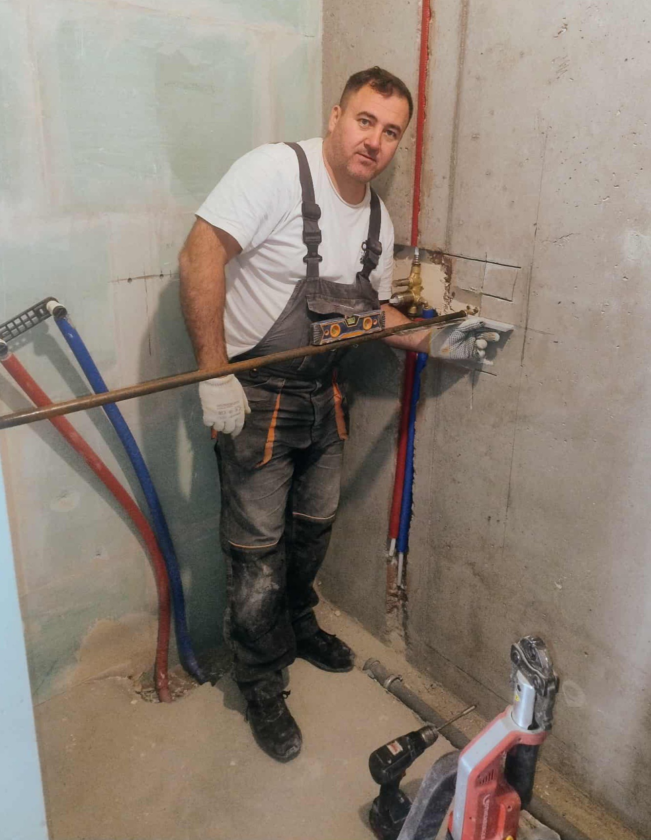 Hydraulik w roboczym kombinezonie, z poziomicą, montuje instalację wodną w surowym wnętrzu. Widoczne narzędzia i rury.