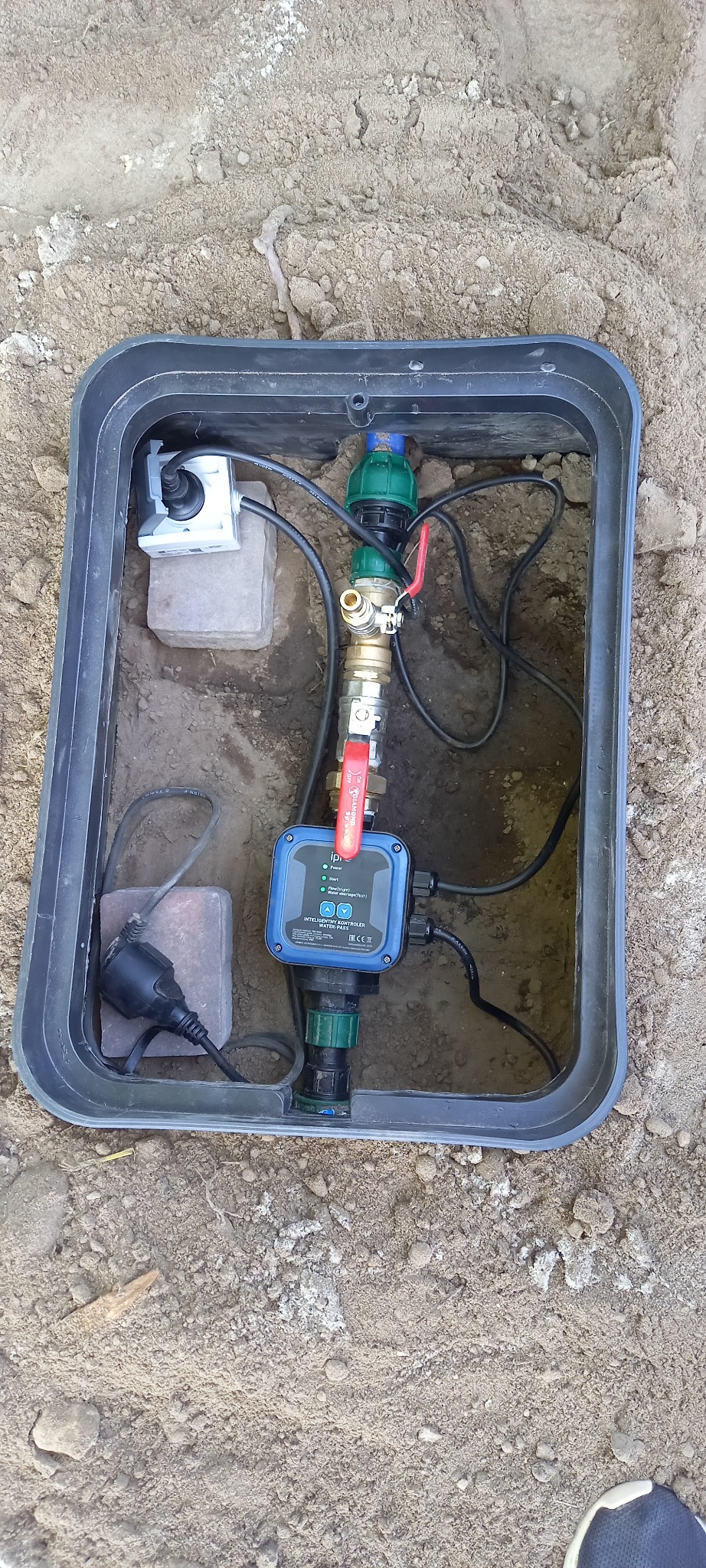 Zewnętrzna skrzynka z instalacją wodną, zawierająca zawory, sterownik ciśnienia i przewody elektryczne, umieszczona w ziemi.