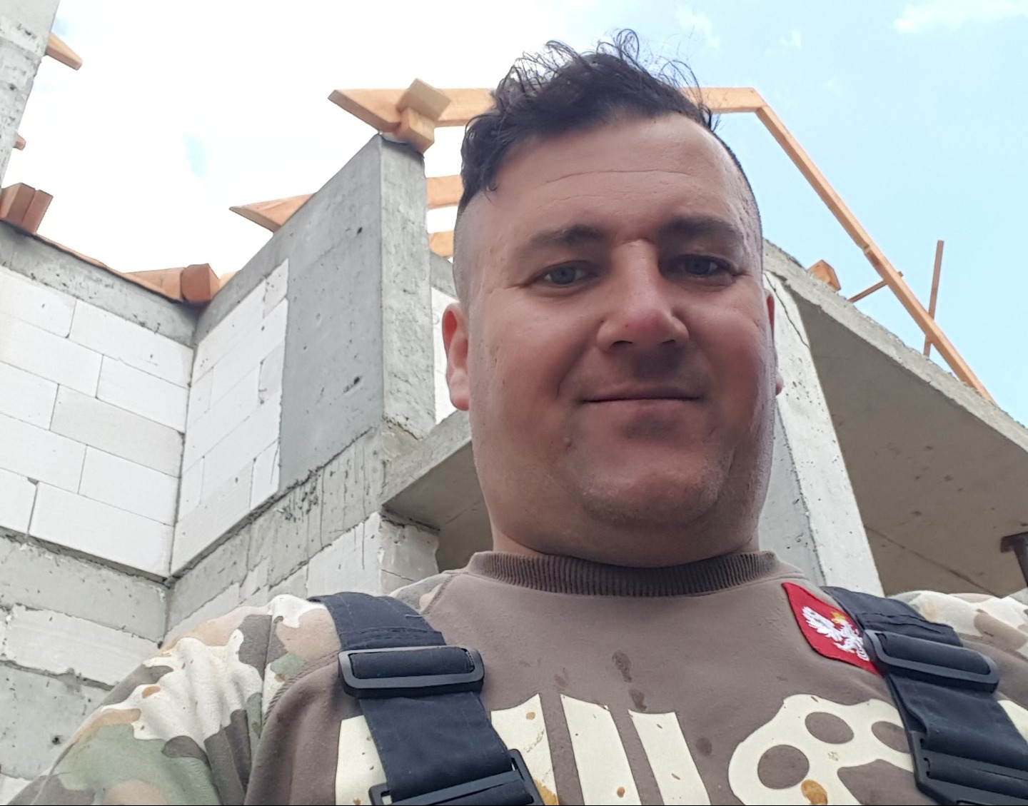 Uśmiechnięty murarz w kombinezonie na tle surowego stanu budynku z betonu komórkowego i drewnianej konstrukcji dachu w trakcie budowy.