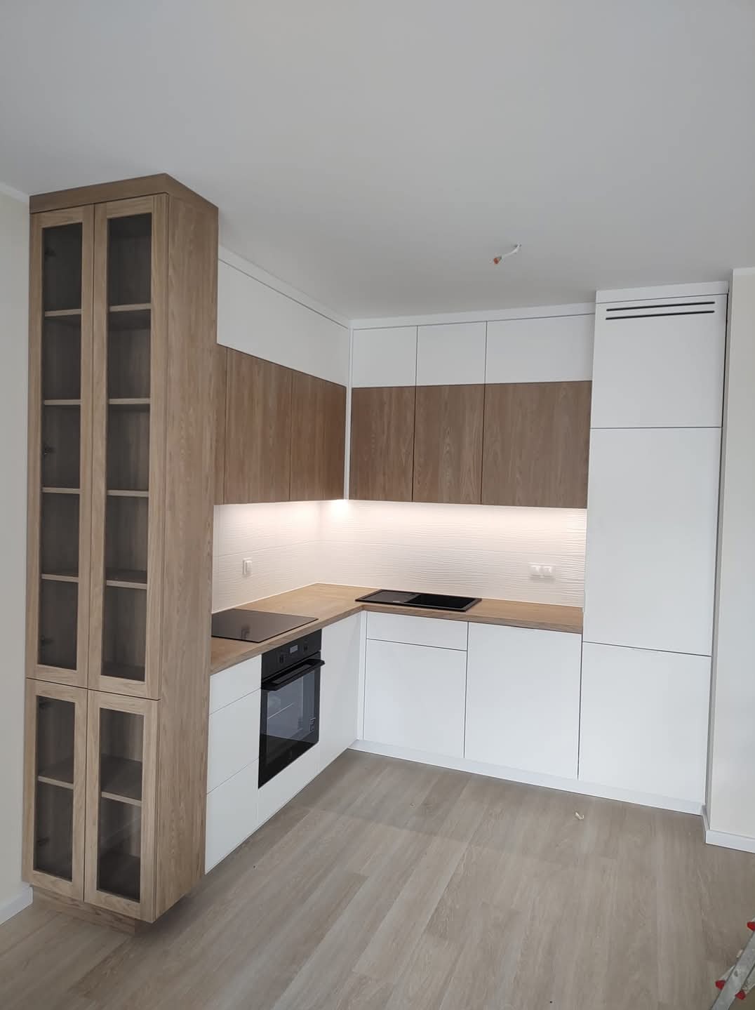 Nowoczesna kuchnia w kształcie litery L z białymi frontami i drewnianymi akcentami. Wysoka szafka z przeszklonymi drzwiami. Oświetlenie LED pod szafkami górnymi.