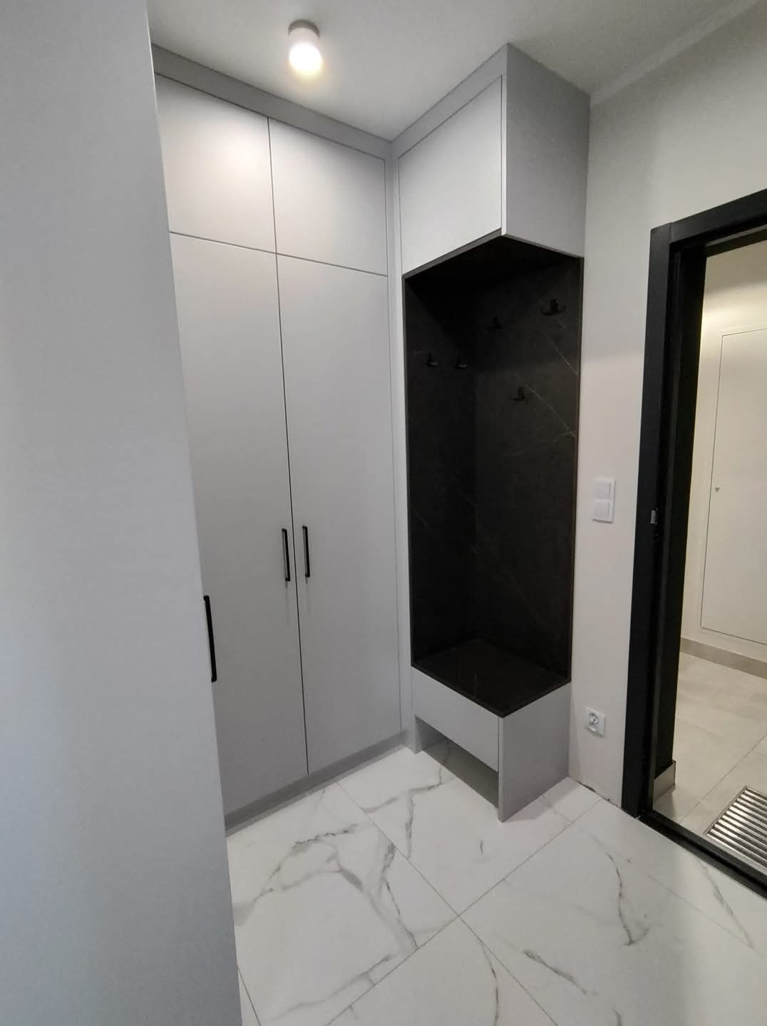 Nowoczesna szafa wnękowa z siedziskiem i wieszakami w przedpokoju. Białe fronty szafy, czarne uchwyty i marmurowe wykończenie wnęki. Minimalistyczny design.