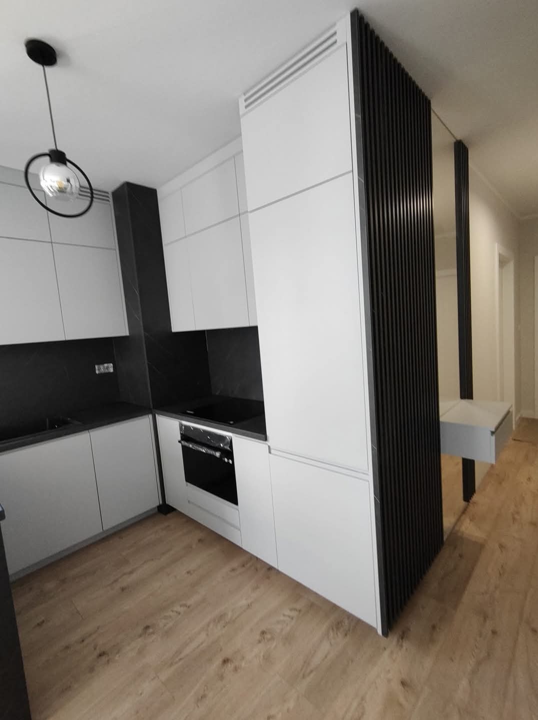 Nowoczesna kuchnia z białymi szafkami i czarnym blatem, oświetlona wiszącą lampą. Na podłodze panele imitujące drewno. Widoczna ażurowa ścianka działowa.