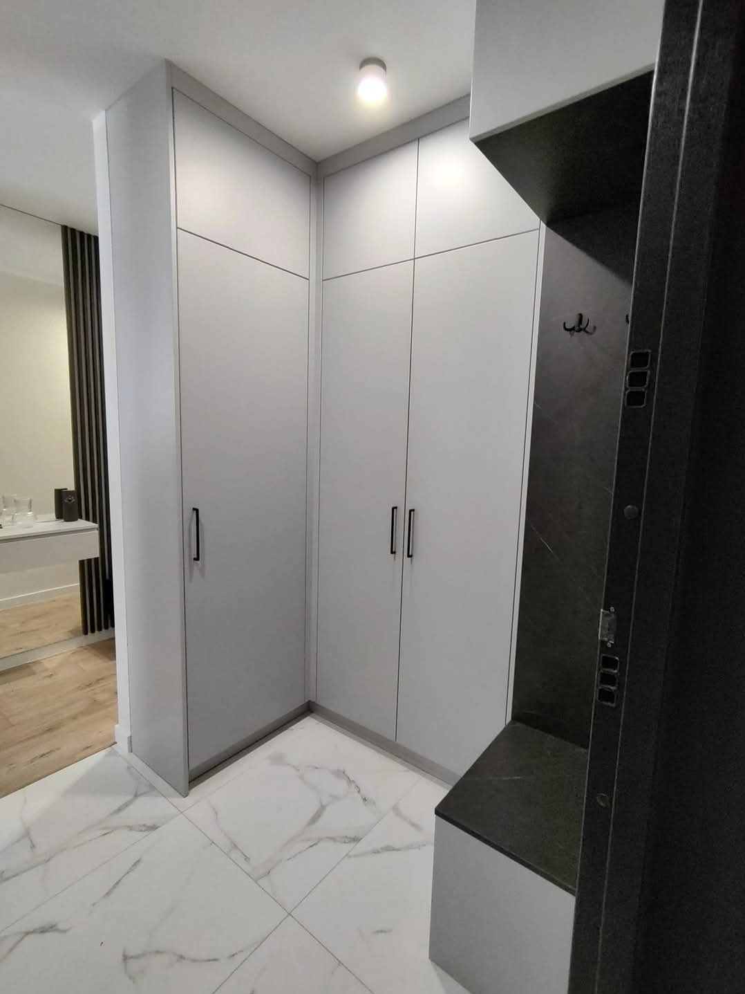 Jasnoszara szafa wnękowa z czarnymi uchwytami, dopasowana do rogu pomieszczenia z marmurową podłogą. Obok wieszak z siedziskiem w ciemnej tonacji.