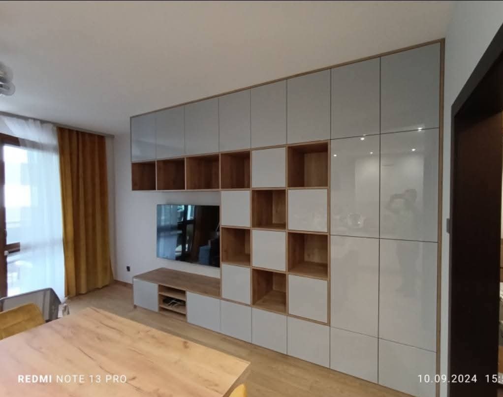 Biała, lakierowana meblościanka na wymiar z drewnianymi wnękami i szafkami, zintegrowana z szafką RTV pod telewizorem. Nowoczesny design.