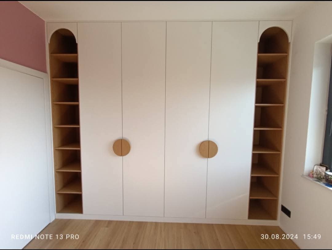 Biała szafa wnękowa z drewnianymi półkami po bokach i okrągłymi uchwytami, widok z przodu na tle jasnych ścian i podłogi. Minimalistyczny design.