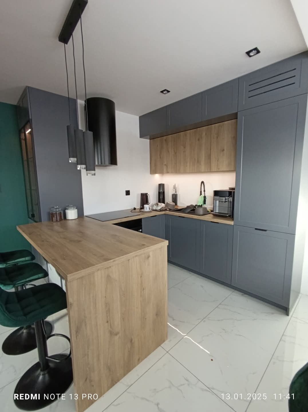 Nowoczesna kuchnia z szarymi szafkami, drewnianym blatem i wyspą. Nad blatem wiszą czarne lampy. Biała podłoga z marmurkowym wzorem. Minimalistyczny design.