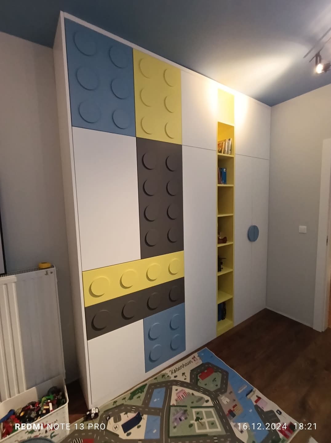 Biała szafa na wymiar z frontami w stylu klocków Lego (niebieski, żółty, szary) w pokoju dziecięcym. Dodatkowa otwarta półka w kolorze żółtym.