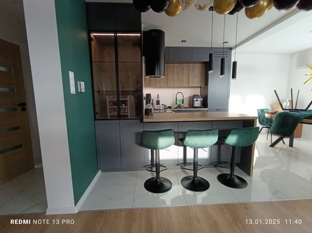 Nowoczesna kuchnia z szarymi szafkami, drewnianym blatem i zielonymi hokerami. Nad blatem wiszą czarne lampy. Dekoracja z balonów dodaje wnętrzu odświętnego charakteru.