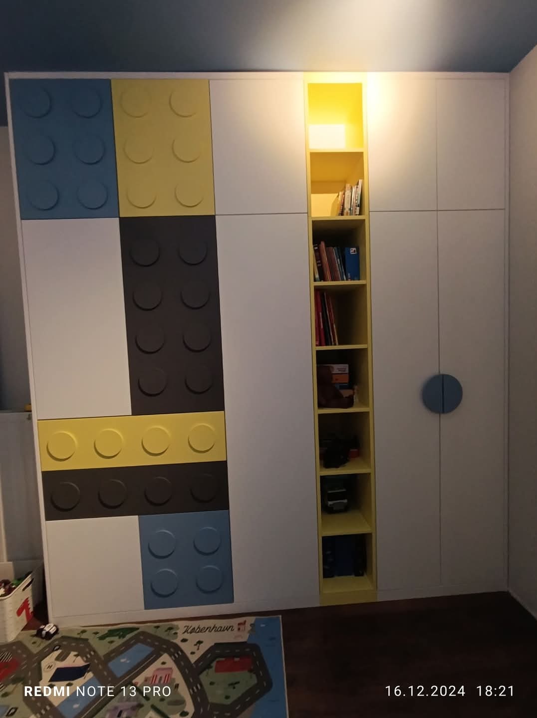 Biała szafa z kolorowymi elementami przypominającymi klocki Lego, z żółtą wnęką na książki i zabawki. Dziecięcy pokój, dywan z motywem miasta.