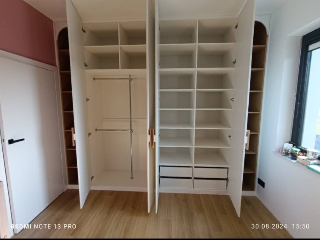 Biała szafa wnękowa z otwartymi drzwiami, widoczne półki i drążki na ubrania. Realizacja w Halinowie. Boczne regały z półkami. Nowoczesny design.