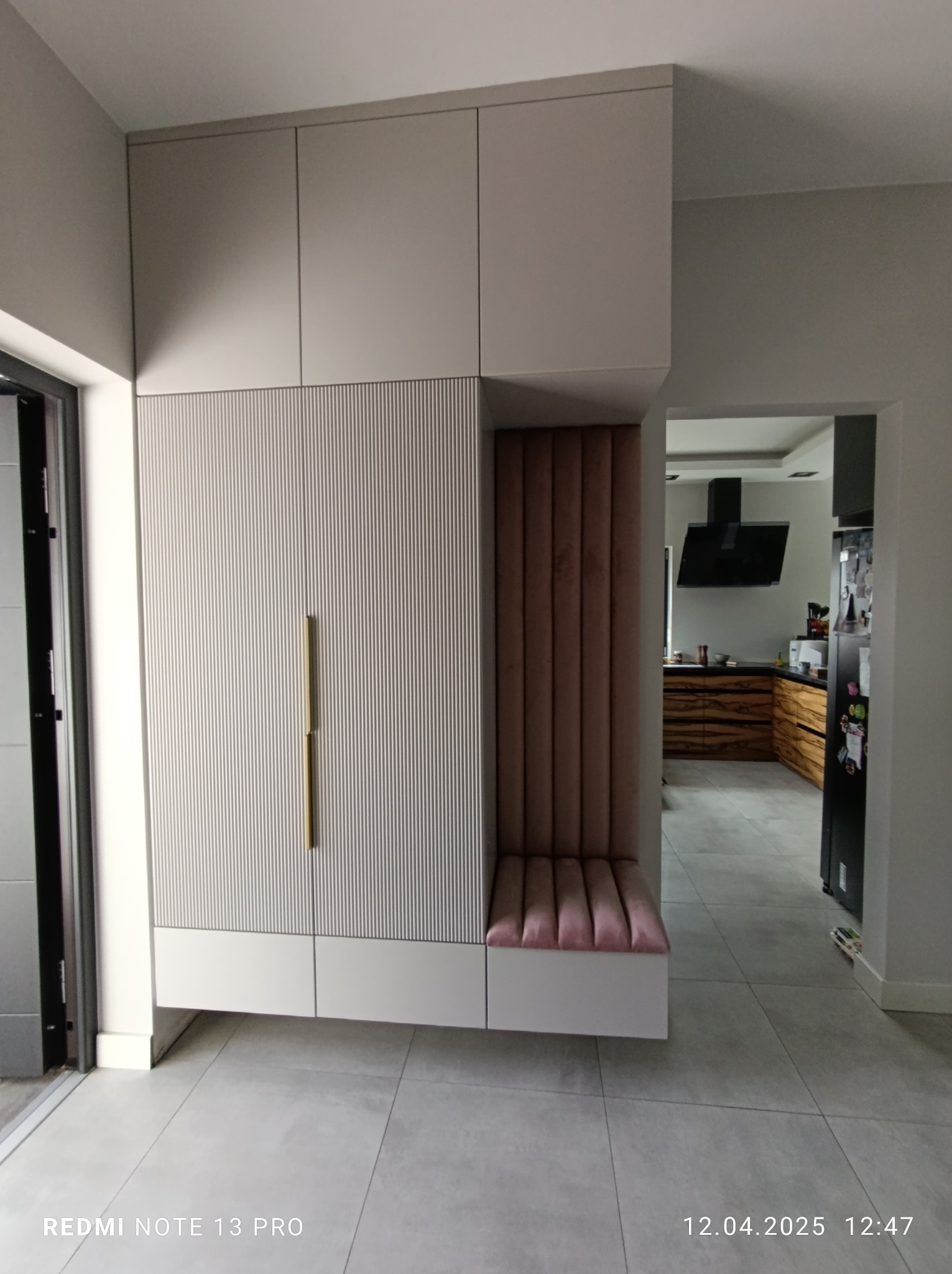 Nowoczesna szafa wnękowa z siedziskiem tapicerowanym różowym welurem, zintegrowana z przedpokojem. Minimalistyczny design, złote uchwyty, widok na kuchnię w tle.