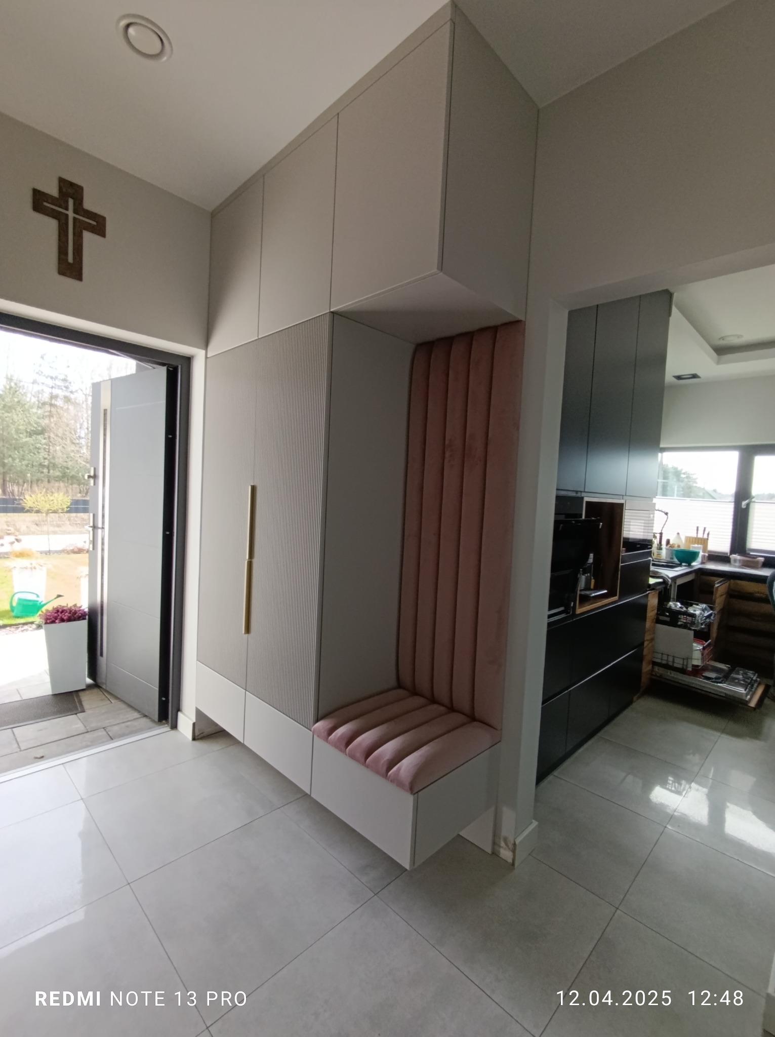 Szafa wnękowa z siedziskiem w przedpokoju, jasne fronty z frezowaniem, złote uchwyty, tapicerowane siedzisko i oparcie w kolorze pudrowego różu. Widok na kuchnię.