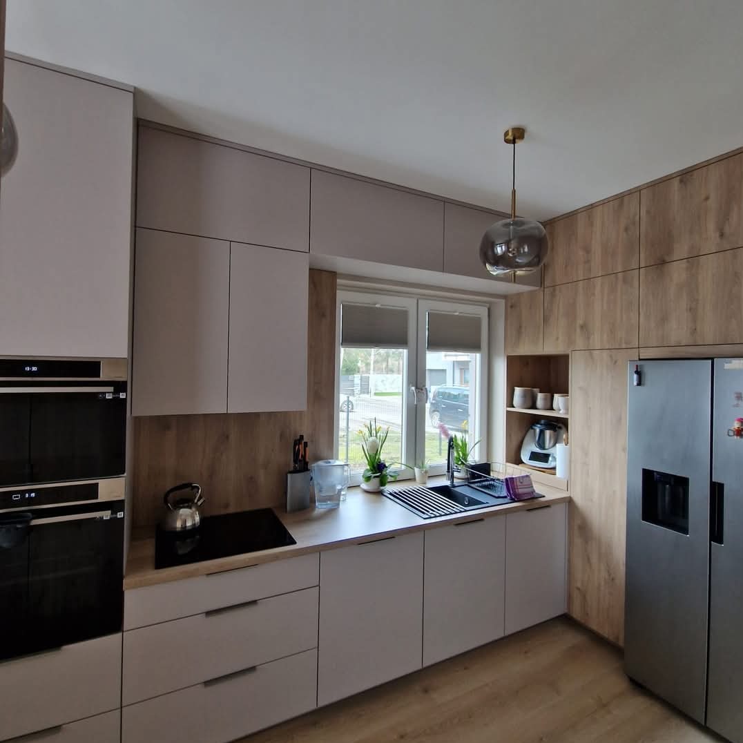 Nowoczesna kuchnia z drewnianymi i jasnymi frontami, oświetlona wiszącą lampą. Widok na okno z roślinami i zabudowaną lodówkę.