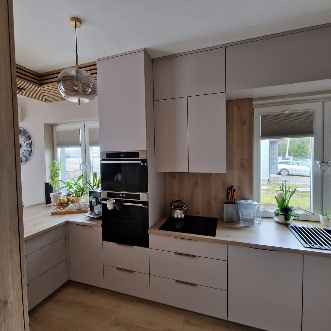 Jasna kuchnia z drewnianymi akcentami: zabudowa w odcieniach szarości, sprzęt AGD w zabudowie, drewniany panel ścienny i blat, lampa wisząca z przezroczystego szkła.