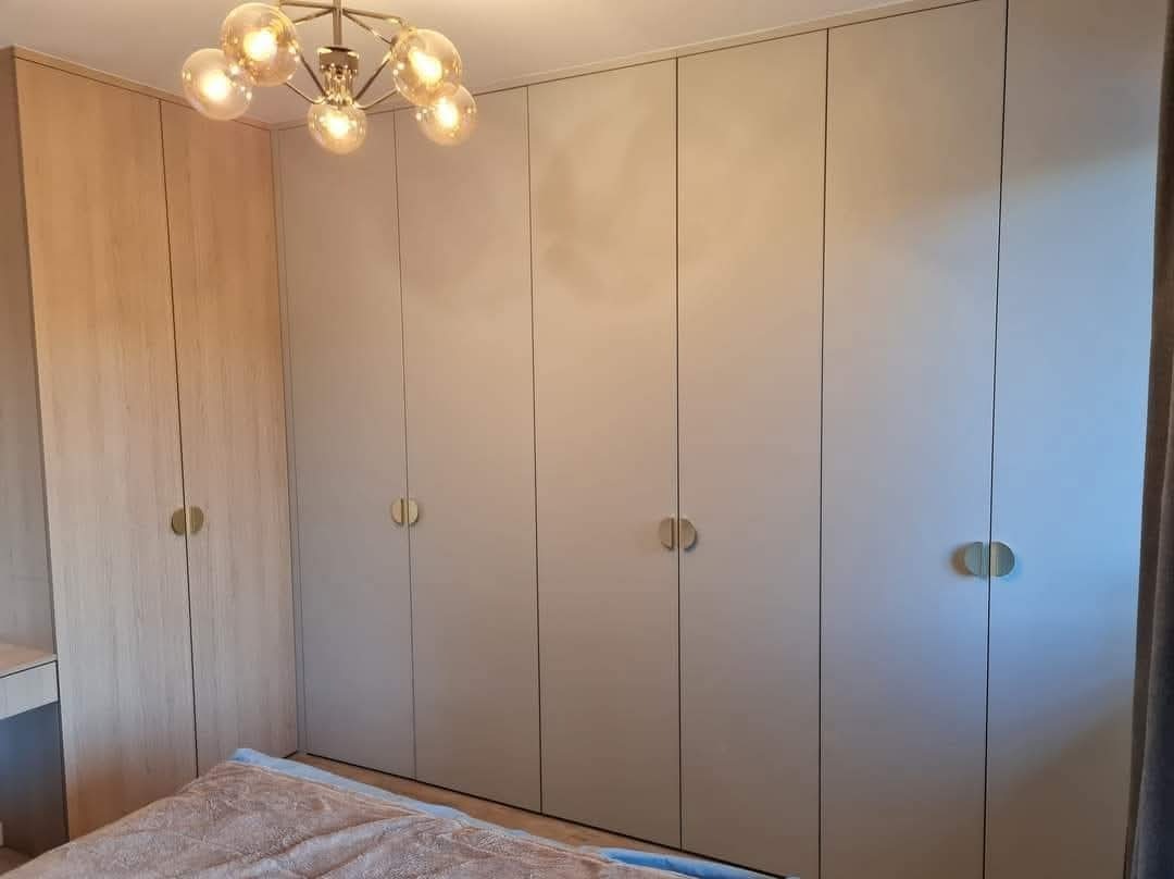 Szafa wnękowa z jasnymi frontami i okrągłymi uchwytami, oświetlona dekoracyjną lampą sufitową. Widoczny fragment łóżka i ściana w tle.