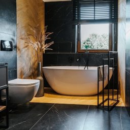 Przemek Wagner - Elegancka łazienka z czarnymi płytkami imitującymi marmur, wolnostojącą białą wanną i drewnianymi akcentami. Nowoczesny design z minimalistycznym wyposażeniem.