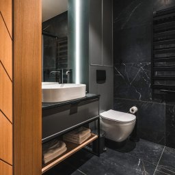 Przemek Wagner - Elegancka, ciemna łazienka z marmurowymi płytkami, owalną umywalką na czarnym blacie i drewnianymi drzwiami z geometrycznym wzorem po lewej stronie.