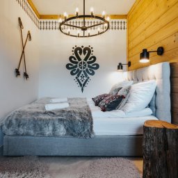 Przemek Wagner - Przytulna sypialnia w góralskim stylu: drewniane panele ścienne, dekoracyjny wzór na ścianie, oryginalne lampy i żyrandol, łóżko z futrzaną narzutą i stolik nocny z pnia.