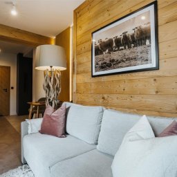 Przemek Wagner - Drewniana ściana z obrazem owiec nad szarą sofą z poduszkami. W tle widoczna kuchnia i drzwi z wzorem w jodełkę. Ciepłe oświetlenie wnętrza.