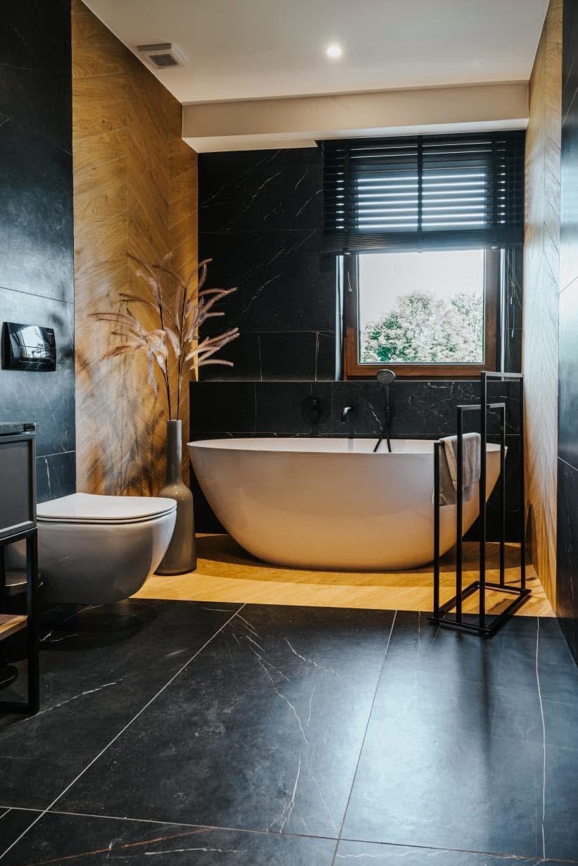 Elegancka łazienka z czarnymi płytkami imitującymi marmur, wolnostojącą białą wanną i drewnianymi akcentami. Nowoczesny design z minimalistycznym wyposażeniem.